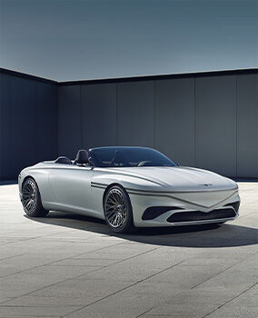The Genesis X Convertible Concept vehicle is parked diagonally with the roof open, situated outdoors and surrounded by gray building walls. The front and right side of the vehicle are visible, with the light gray bodywork appearing glossy in the sunlight.